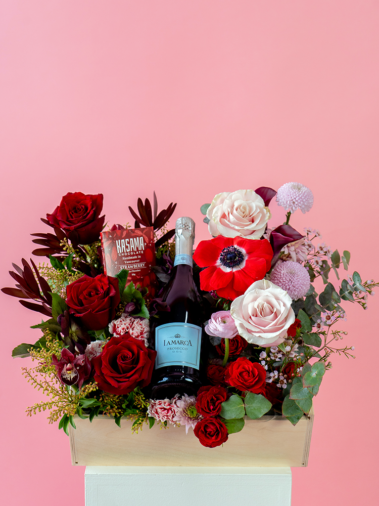 Vancouver Florist - Vancouver Flower Delivery - Valentine's Flowers, A floral gift box with a bottle of prosecco and various flowers including red roses, pink flowers, and greenery, presented on a wooden surface with a pink background.
